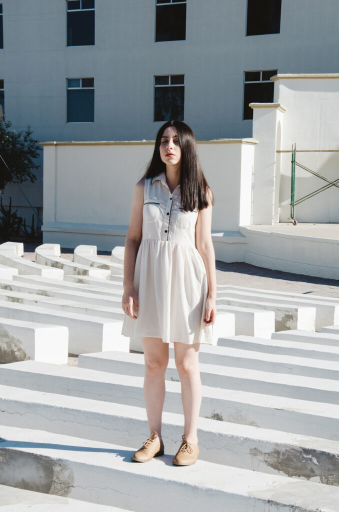 Gabriela Inzunza stands on white steps amidst a sunlit courtyard wearing a white dress and brown shoes with a backdrop of a white building with windows.